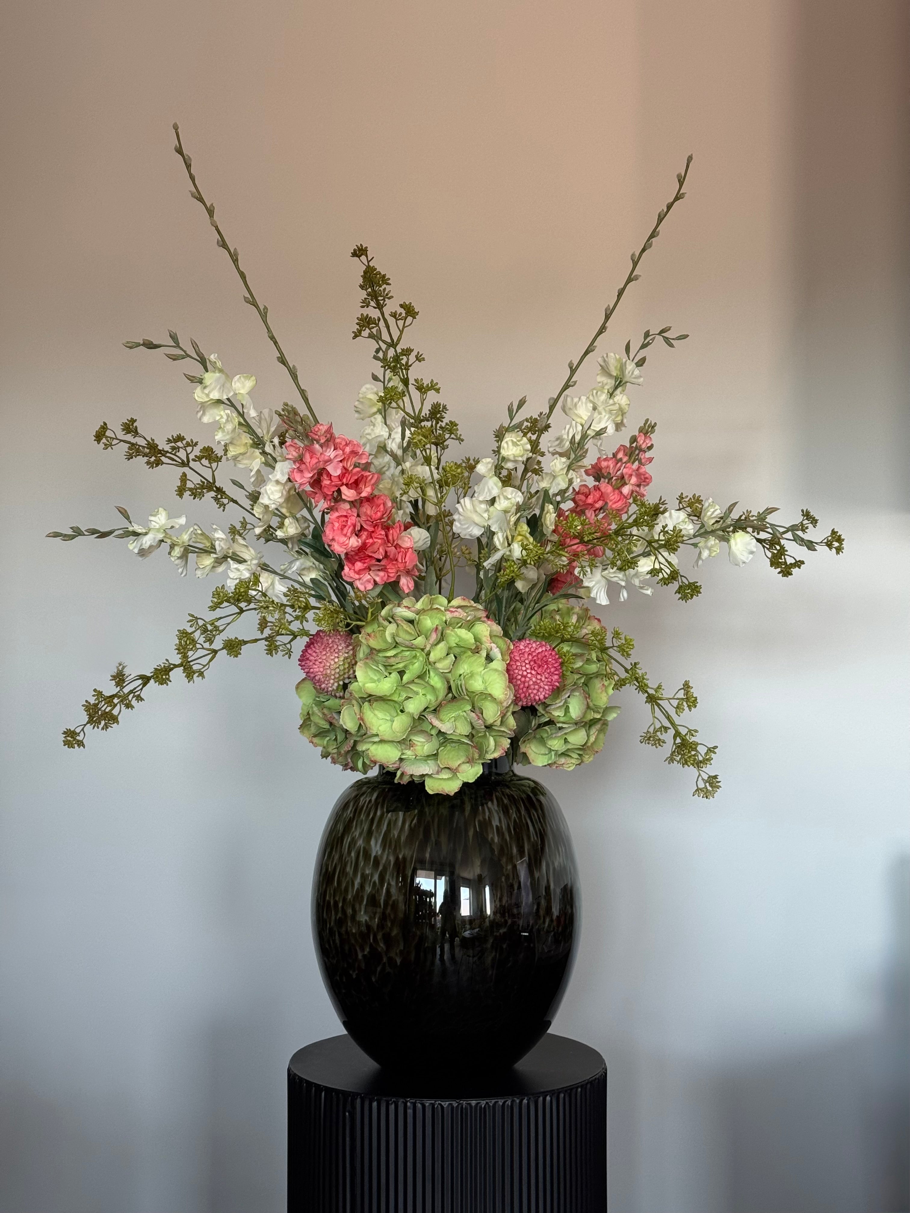 Groot zijden boeket met groene hortensia's en roze bloemen in een unieke mondgeblazen zwarte vaas op een zwarte sokkel.
